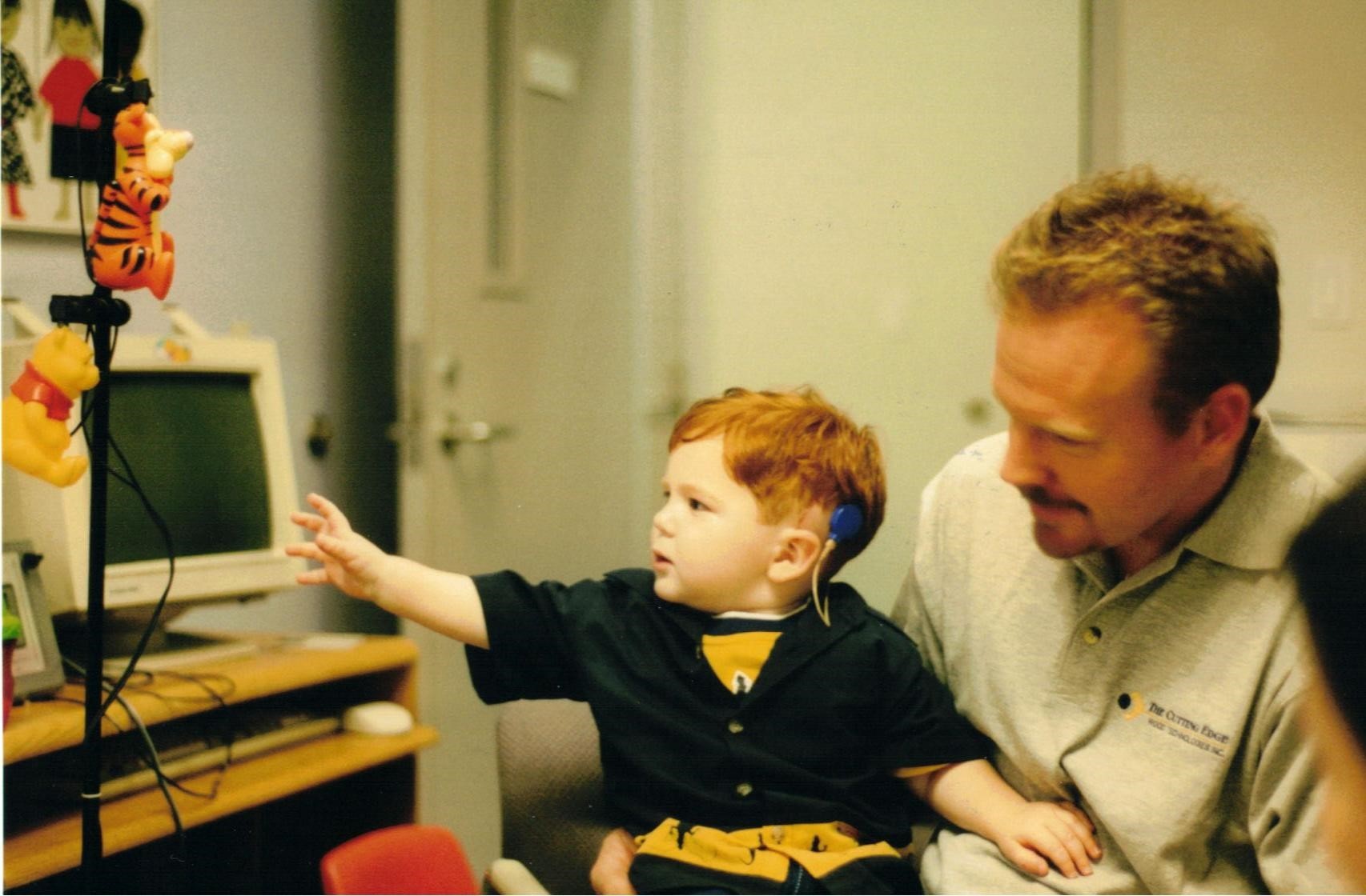 A picture of young Matthew sitting on a man's lap during his cochlear implant journey.