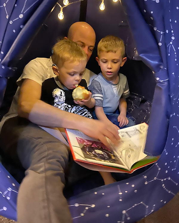 a father reads a book to his young sons, one of whom has has a cochlear implant